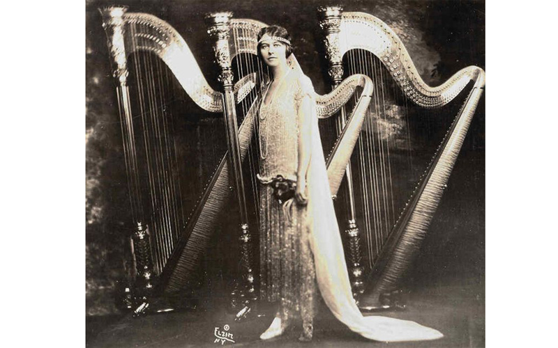 In an old photograph, a bride in a wedding dress and a bouquet stands next to a tar instrument.